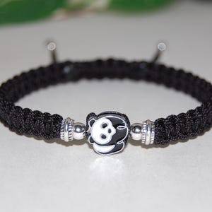 Puede incluir: Pulsera trenzada negra con un dije plateado con un panda blanco y negro.