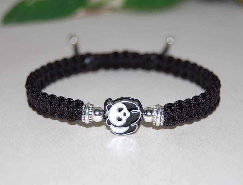 Puede incluir: Pulsera trenzada negra con un dije plateado con un panda blanco y negro.