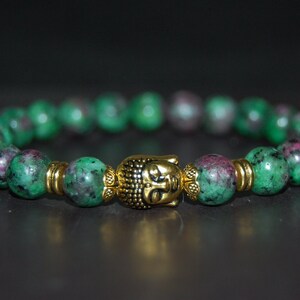 May include: A green and pink gemstone bracelet with a gold Buddha charm. The bracelet is made of round beads and has a simple design.