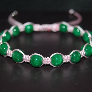 Puede incluir: Un brazalete de piedras verdes con cordón rosa. El brazalete está hecho con cuentas redondas verdes y un cordón rosa anudado.