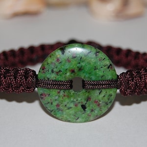May include: A green and brown bracelet with a round green stone with black and red flecks. The stone has a hole in the center and is surrounded by brown cord.