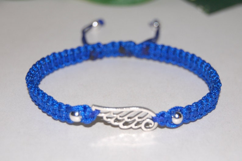 May include: A blue macrame bracelet with a silver wing charm. The bracelet is adjustable with sliding knots and features silver beads at each end. The wing charm is centered on the bracelet.