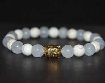 Op de afbeelding: Een armband met afwisselende witte en lichtblauwe kralen, met een gouden Boeddha-kop bedel.