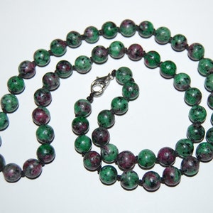 May include: A green and pink gemstone necklace with a silver clasp. The necklace is made of round beads and is strung on a black cord.