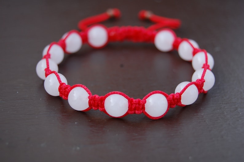 May include: A red string bracelet with white round beads. The bracelet is tied with a knot.