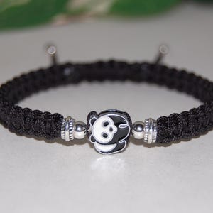 Puede incluir: Pulsera trenzada negra con un dije plateado con dise&ntilde;o de oso panda.