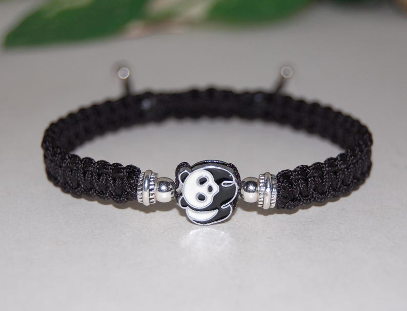 Puede incluir: Pulsera trenzada negra con un dije plateado con dise&ntilde;o de oso panda.