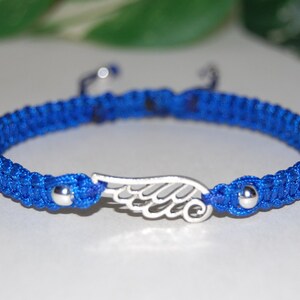 May include: A blue macrame bracelet with a silver wing charm. The bracelet features a woven design with a sliding knot closure. The wing charm is centered on the bracelet and has intricate detailing. The bracelet is approximately 7 inches in length.