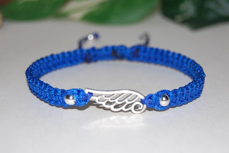 May include: A blue macrame bracelet with a silver wing charm. The bracelet features a woven design with a sliding knot closure. The wing charm is centered on the bracelet and has intricate detailing. The bracelet is approximately 7 inches in length.
