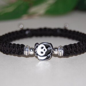 Puede incluir: Pulsera trenzada negra con un dije plateado con un dise&ntilde;o de panda blanco.