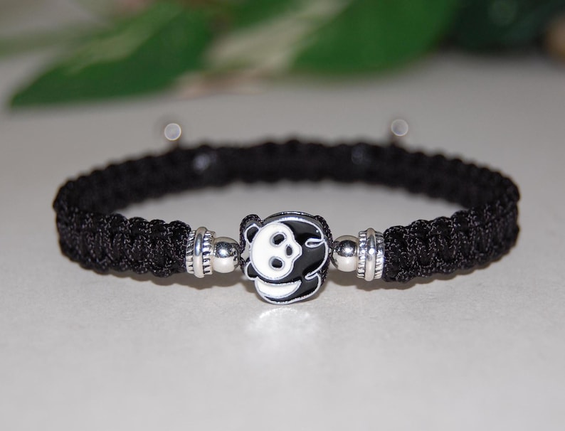 Puede incluir: Pulsera trenzada negra con un dije plateado con un dise&ntilde;o de panda blanco.