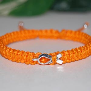 Könnte beinhalten: Orangefarbenes geflochtenes Armband mit einem silbernen Anhänger in Form eines Bandes mit der Aufschrift "HOPE".