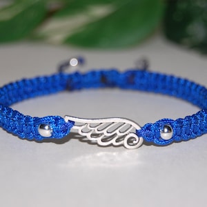 May include: A blue macrame bracelet with a silver wing charm. The bracelet features a woven design with a sliding knot closure. The wing charm is centered on the bracelet and has intricate detailing. The bracelet is a vibrant blue color.