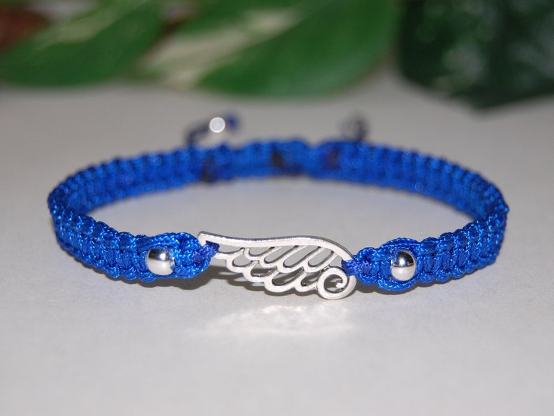 May include: A blue macrame bracelet with a silver wing charm. The bracelet features a woven design with a sliding knot closure. The wing charm is centered on the bracelet and has intricate detailing. The bracelet is a vibrant blue color.