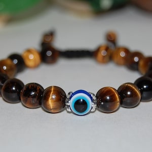 May include: A beaded bracelet featuring tiger's eye stones and a central blue evil eye bead. The bracelet's beads are a mix of brown and gold, strung on a black cord. The evil eye bead is surrounded by silver-colored accents.