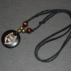 Può includere: Una collana con cordoncino nero con un ciondolo rotondo in legno che presenta un design di lupo bianco. La collana ha perline di legno marrone e accenti metallici dorati.