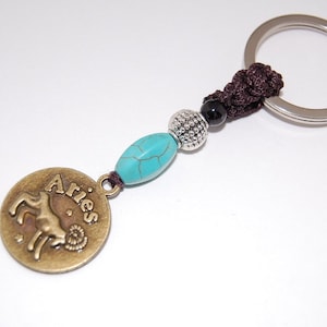 May include: A turquoise and black beaded keychain with a silver charm featuring the zodiac sign Aries and the word "Aries".