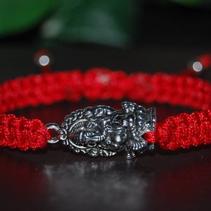 Peut inclure: Bracelet tressé rouge avec un charme argenté orné d'un motif détaillé.