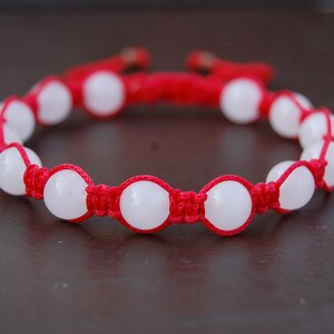 May include: A red string bracelet with white round beads. The bracelet is knotted with a simple design.