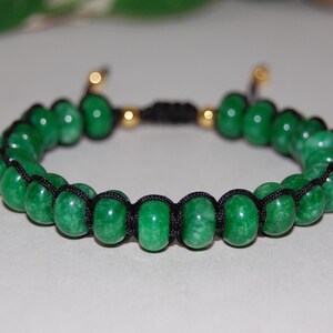 Puede incluir: Un brazalete de piedra de jade verde con cordón negro y detalles dorados. El brazalete está hecho de cuentas redondas y tiene un cierre de nudo deslizante.
