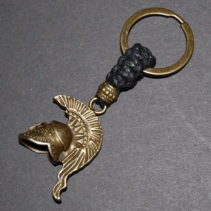 May include: A gold-toned key chain featuring a detailed Roman helmet charm and a black braided cord. The key chain has a circular ring for attaching keys.