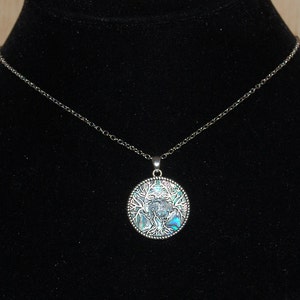 May include: A silver necklace with a circular pendant depicting a tree. The pendant has iridescent blue and green details. The necklace is shown against a black background.