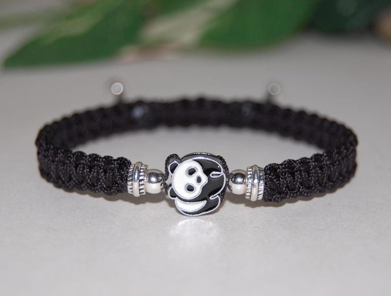 Puede incluir: Pulsera trenzada negra con un dije de panda plateado. El panda es negro y blanco con un contorno negro.