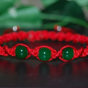 Puede incluir: Un brazalete de cuerda roja con tres cuentas verdes. El brazalete está hecho de un cordón trenzado y las cuentas son redondas y lisas.