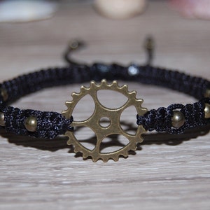 Könnte beinhalten: Ein schwarzes geflochtenes Armband mit einem bronzenen Zahnrad-Anhänger und kleinen bronzenen Perlen.