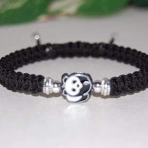 Puede incluir: Pulsera trenzada negra con un dije plateado con la cara de un panda.