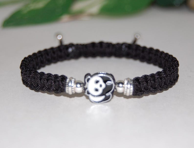 Puede incluir: Pulsera trenzada negra con un dije plateado con la cara de un panda.