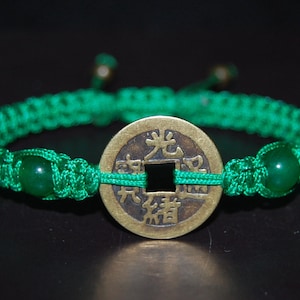 Puede incluir: Un brazalete trenzado verde con un dije redondo de moneda de color latón con caracteres chinos. El brazalete también tiene dos cuentas de jade verde.