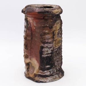 Może przedstawiać: Cylindryczny ceramiczny wazon o teksturowanej powierzchni. Wazon ma paletę kolorów brązowego, czerwonego i beżowego. Górna krawędź jest ciemnobrązowa, a korpus ma szorstką, nierówną fakturę z wypukłymi detalami.