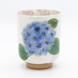 Può includere: Una tazza da tè in ceramica bianca con un design di fiori di ortensia blu e dorati. La tazza ha un bordo e una base marroni.