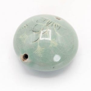 May include: A round, pale green ceramic bead with a glossy, crackled glaze finish. The surface features a delicate, etched botanical design in white. The bead has two visible circular holes, one on the side and one near the top edge.