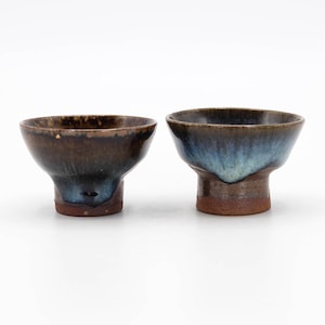 May include: Two small ceramic teacups with brown and blue glaze. The cups have a flared lip and a small foot.