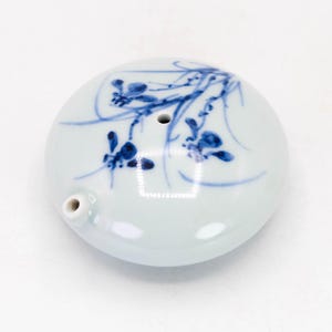 Puede incluir: Un tintero de cerámica blanca con un diseño floral azul y blanco. El tintero tiene un pequeño pico y un agujero en la parte superior para sumergir un pincel.