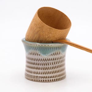 Puede incluir: Un recipiente de cerámica para utensilios con borde esmaltado azul y blanco y un patrón texturizado. Una cuchara de madera y una taza de bambú descansan dentro del recipiente, creando una estética natural y rústica. Los artículos están sobre un fondo blanco.