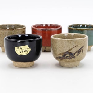 Könnte beinhalten: Fünf Keramik-Teetassen in verschiedenen Farben und Designs. Die Tassen sind alle klein und haben eine abgerundete Form. Eine Tasse ist schwarz mit einem goldenen Etikett, auf dem "民芸 からつ焼" steht.