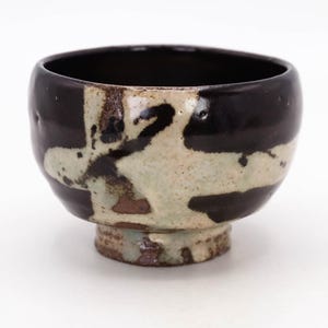 May include: A handcrafted ceramic bowl with a dark brown exterior and a light beige and brown patterned design. The bowl has a rounded shape with a small, pedestal base. The interior is a glossy dark brown color. This unique bowl is perfect for serving soups or other dishes.