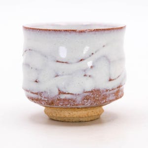 May include: A handmade ceramic tea bowl with a white glaze and textured surface. The bowl has a reddish-brown base and rim, and sits on a small, unglazed foot. The design is simple and elegant, suitable for serving tea or other beverages.