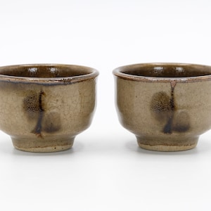 Puede incluir: Dos pequeñas tazas de té de cerámica marrón con un esmalte marrón oscuro y un sutil acabado craquelado. Las tazas tienen una forma redondeada y un labio ligeramente acampanado.