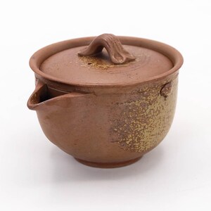 Puede incluir: Una tetera de cerámica marrón con tapa y asa. La tetera tiene un pico y una superficie texturizada. La tapa tiene un asa pequeña. La tetera está diseñada para preparar té.
