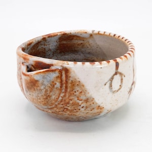 Puede incluir: Un pequeño cuenco de cerámica blanca con esmalte moteado marrón y gris. El cuenco tiene una forma redondeada y un borde ligeramente acampanado. Tiene un pequeño pico en el lateral.