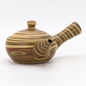 May include: A striped teapot with a lid and a curved handle. The teapot features a pattern of alternating bands of brown, tan, and red. The spout is curved and angled. The teapot is made of ceramic or a similar material.