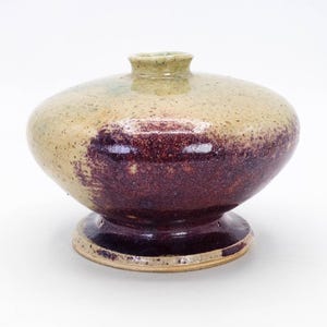 May include: A small, round ceramic vase with a speckled glaze in shades of beige, green, and purple. The vase has a wide base and a narrow neck.