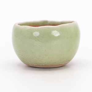Può includere: Una piccola ciotola in ceramica rotonda con un esterno smaltato verde menta chiaro e una sottile finitura craquelé. Il bordo ha una forma organica, morbida e ondulata, e la base rivela il materiale naturale in argilla bruno-rossastra.
