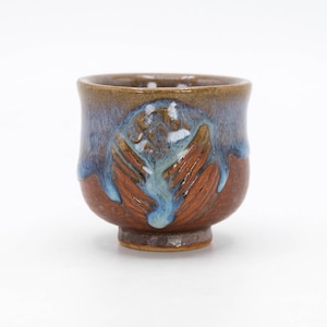 May include: A small, brown ceramic cup with a blue and brown glaze. The cup has a unique, textured design and a rounded base.