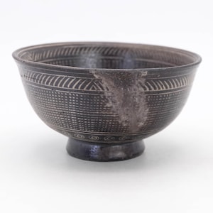 May include: A dark gray ceramic bowl with a pedestal base. The bowl features intricate etched patterns, including geometric shapes and lines. The design is in a lighter shade of gray, creating a contrast against the darker background. The bowl is likely used for serving or display.