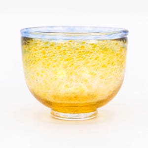 May include: A handmade glass bowl with a rounded form. The bowl has a speckled yellow and orange pattern, with a light blue rim. The glass is transparent. The bowl sits on a clear base. The bowl's measurements are in centimetres.